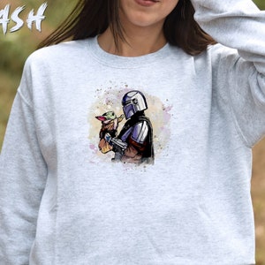 May include: A gray sweatshirt with a watercolor illustration of a Mandalorian character holding a small green alien creature. The illustration is in a light blue, purple, and green color scheme.