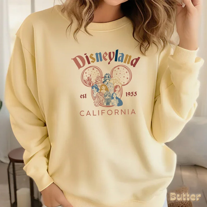 May include: A yellow sweatshirt with a vintage-style Disneyland logo featuring a Mickey Mouse head with a castle and Disney princesses inside. The text reads "Disneyland est 1955 California".