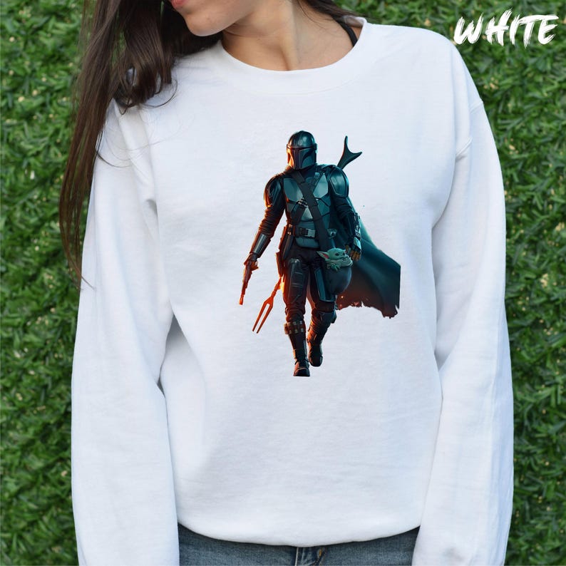 May include: White long-sleeved sweatshirt featuring a printed image of a Mandalorian character from Star Wars. The character is wearing a helmet and holding a weapon. The character is standing in front of a blurred background.