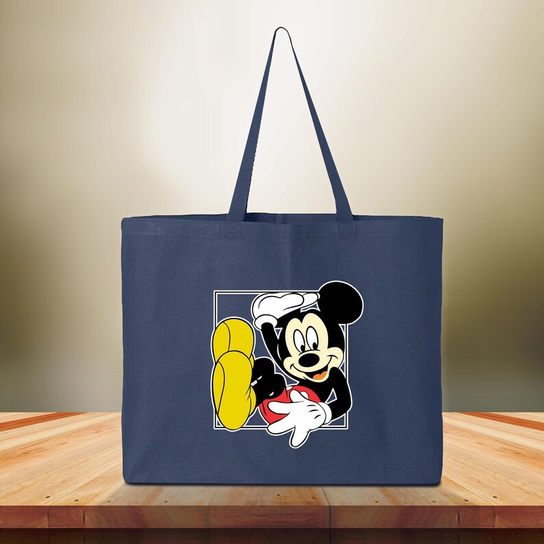 May include: Navy blue tote bag featuring a graphic of Mickey Mouse in a white square. The cartoon character is black, white, red, and yellow. The bag has two navy blue handles and is made of a sturdy fabric.
