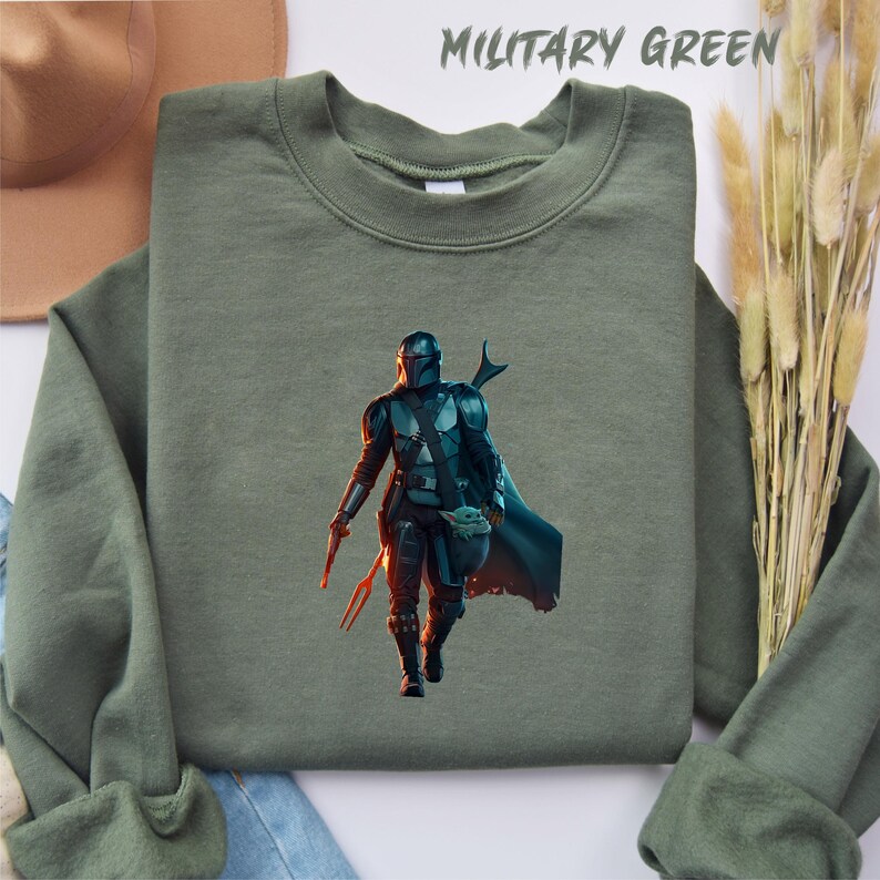 May include: A green sweatshirt featuring a printed image of a character from the Star Wars series, The Mandalorian, holding a small creature. The text "Military Green" is printed in white at the top of the image.