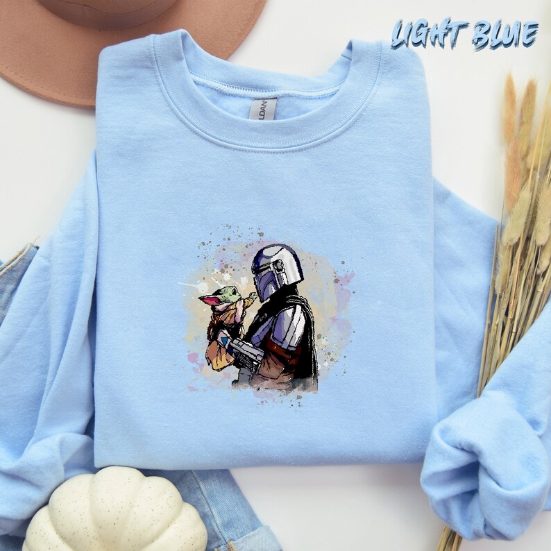 May include: Light blue sweatshirt with a watercolor illustration of a Mandalorian character holding a small green alien creature. The illustration is surrounded by a light blue background with white splatters.