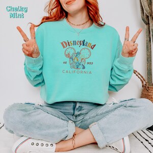 May include: A turquoise blue sweatshirt with a vintage-style Disneyland graphic. The graphic features a Mickey Mouse head with a colourful illustration of Disneyland characters inside. The text "Disneyland" is in a rainbow font with the words "est. 1955" and "CALIFORNIA" below the graphic.