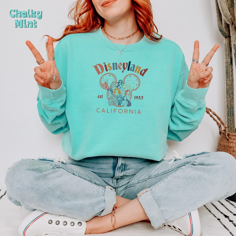 May include: A turquoise blue sweatshirt with a vintage-style Disneyland graphic. The graphic features a Mickey Mouse head with a colourful illustration of Disneyland characters inside. The text "Disneyland" is in a rainbow font with the words "est. 1955" and "CALIFORNIA" below the graphic.