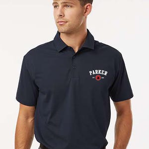Puede incluir: Polo azul marino con cuello y mangas cortas. La camisa presenta la palabra "PARKER" en blanco sobre un dise&ntilde;o circular rojo y blanco. La palabra "navy" est&aacute; en la esquina superior izquierda.