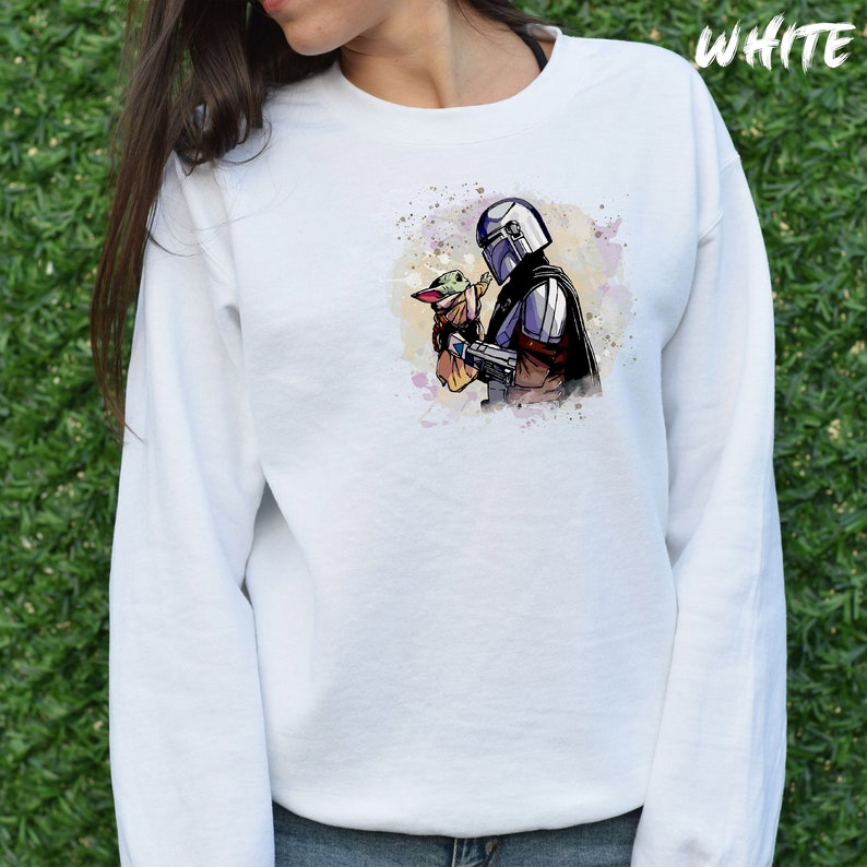 May include: White sweatshirt with a watercolor illustration of a Mandalorian character holding a small green alien character. The illustration is on the front of the sweatshirt.