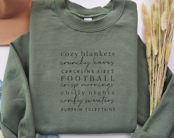 May include: Military green sweatshirt with the words "cozy blankets, crunchy leaves, crackling fires, football, crisp mornings, chilly nights, comfy sweaters, pumpkin everything" in black script. A tan hat and dried flowers are in the background.
