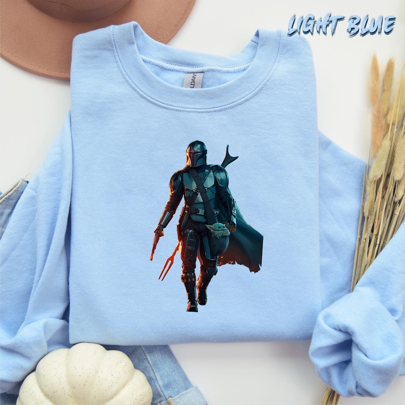 May include: A light blue sweatshirt featuring a graphic print of a Mandalorian character from the Star Wars series. The character is wearing a helmet and holding a weapon. The character is standing in front of a red and orange background.