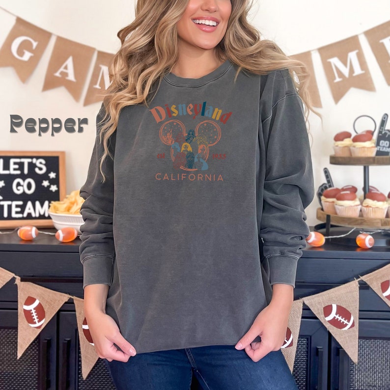 May include: A dark grey sweatshirt with a vintage-style Disneyland logo featuring Mickey Mouse and the text "Disneyland 1955 CALIFORNIA".