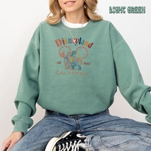 May include: A light green sweatshirt with a vintage-style Disneyland logo featuring Mickey Mouse's head with the text "Disneyland est 1955 CALIFORNIA".