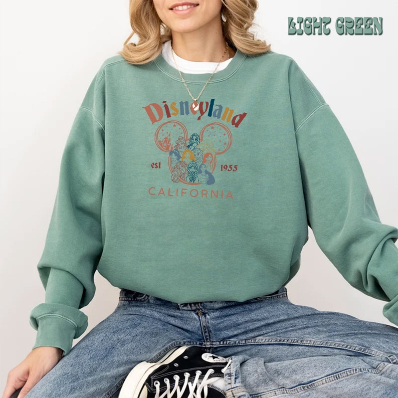 May include: A light green sweatshirt with a vintage-style Disneyland logo featuring Mickey Mouse's head with the text "Disneyland est 1955 CALIFORNIA".