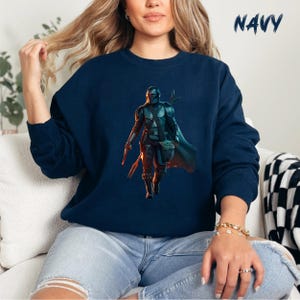 May include: A navy blue sweatshirt featuring a graphic of a Mandalorian character from the Star Wars universe. The character is wearing a helmet and holding a weapon. The character is standing in front of a green alien creature.