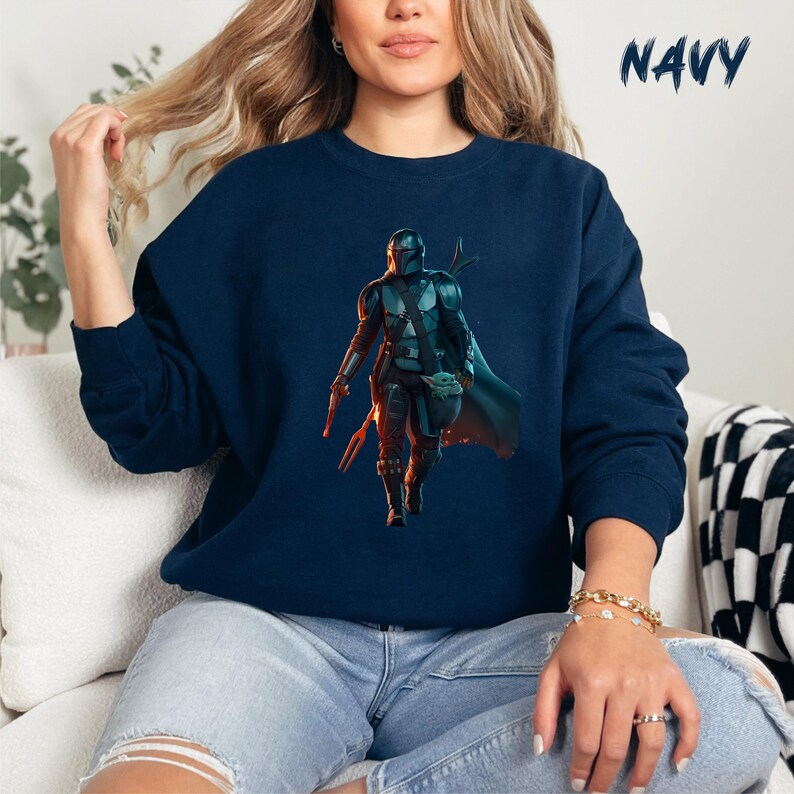 May include: A navy blue sweatshirt featuring a graphic of a Mandalorian character from the Star Wars universe. The character is wearing a helmet and holding a weapon. The character is standing in front of a green alien creature.