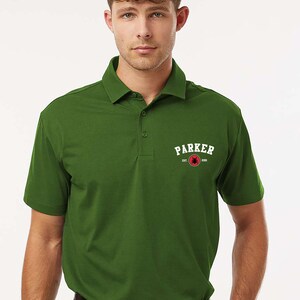 Puede incluir: Polo verde oliva con cuello y mangas cortas. La camisa presenta la palabra "PARKER" en letras blancas con un gr&aacute;fico rojo y negro. El texto "EST. 2005" est&aacute; debajo de la palabra "PARKER".