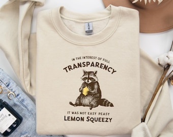 May include: A beige sweatshirt with a raccoon graphic and the text "In the interest of full transparency, it was not easy peasy lemon squeezy."
