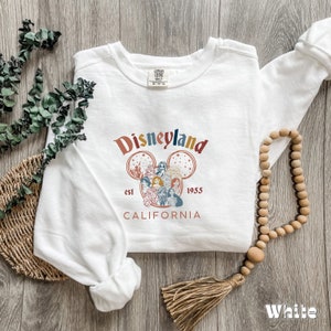 May include: White long-sleeved sweatshirt with a vintage-style Disneyland graphic featuring Mickey Mouse ears, the Disneyland castle, and various Disney characters. The text "Disneyland est 1955 California" is printed below the graphic.