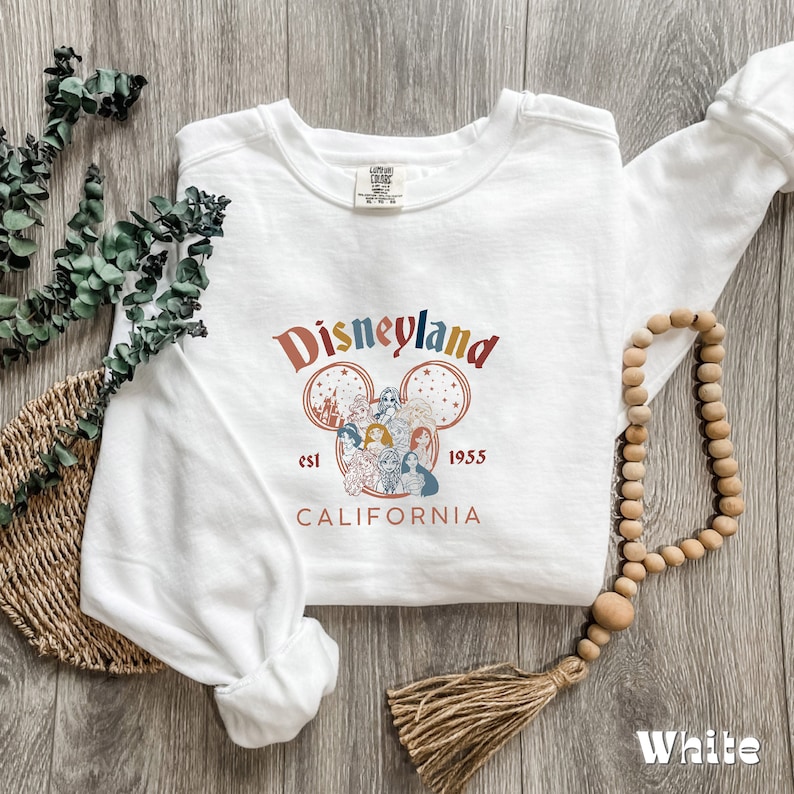 May include: White long-sleeved sweatshirt with a vintage-style Disneyland graphic featuring Mickey Mouse ears, the Disneyland castle, and various Disney characters. The text "Disneyland est 1955 California" is printed below the graphic.