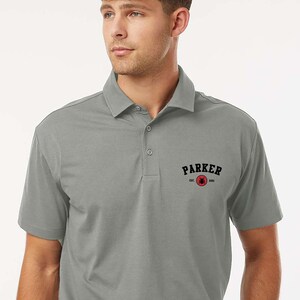Puede incluir: Un polo gris con cuello y mangas cortas. La camisa lleva la palabra "PARKER" en letras negras sobre un logotipo circular rojo. La palabra "greystone" est&aacute; en la esquina superior izquierda.