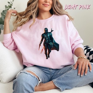 May include: A light pink sweatshirt featuring a graphic print of a Mandalorian character from the Star Wars series. The character is wearing a helmet and holding a small creature in his hand.