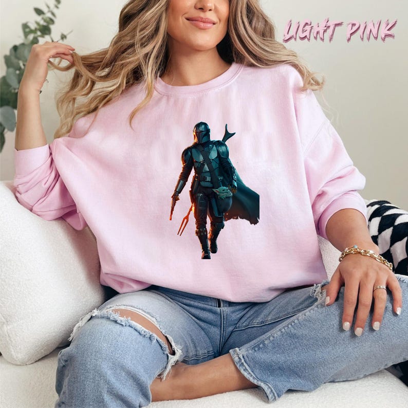 May include: A light pink sweatshirt featuring a graphic print of a Mandalorian character from the Star Wars series. The character is wearing a helmet and holding a small creature in his hand.