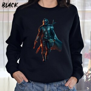 May include: Black sweatshirt featuring a printed image of a Mandalorian character from Star Wars. The character is holding a small, green creature. The text "BLACK" is printed in white in the top left corner of the sweatshirt.