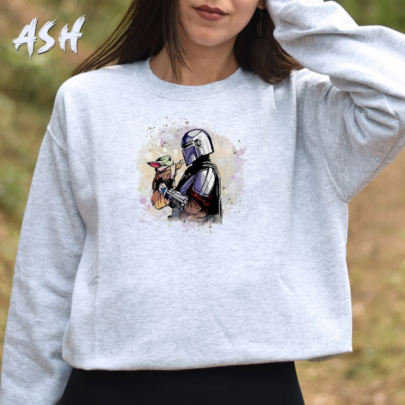 May include: A gray sweatshirt with a watercolor illustration of a Mandalorian character holding a small green alien creature. The illustration is in a light blue, purple, and green color scheme.