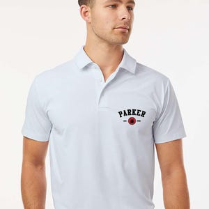 Puede incluir: Polo blanco con cuello y mangas cortas. La camisa presenta un logotipo negro y rojo que dice "PARKER" con "EST. 2023". El modelo lleva pantalones negros y un cintur&oacute;n negro.