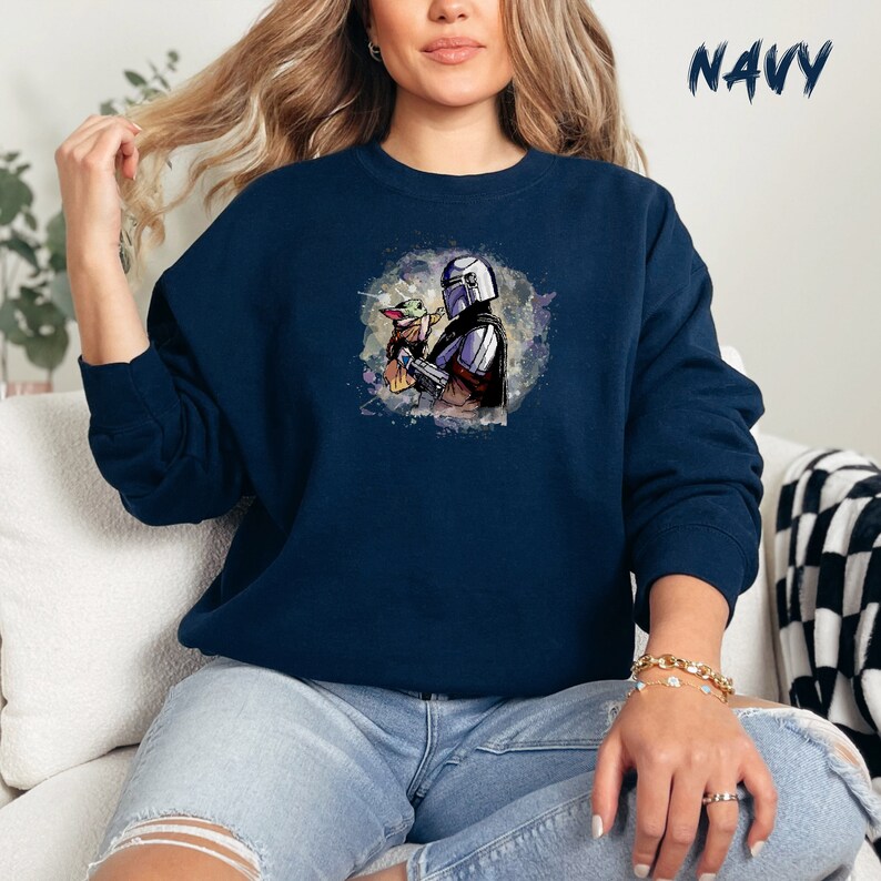 May include: A navy blue sweatshirt with a watercolor illustration of a Mandalorian character holding a small green creature. The illustration is surrounded by a light blue and white watercolor background.