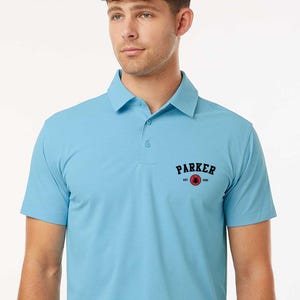 Puede incluir: Polo de color azul claro con cuello y mangas cortas. La camisa tiene la palabra "PARKER" en letras negras con un gr&aacute;fico rojo y negro. El texto "est. 2001" est&aacute; debajo de la palabra "PARKER".