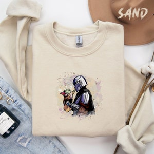 May include: A beige sweatshirt with a watercolor-style illustration of a Mandalorian character holding a small green alien creature. The illustration is in shades of blue, green, and purple.