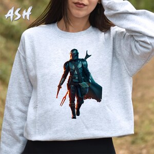 May include: A grey sweatshirt featuring a printed image of a Mandalorian character from the Star Wars franchise. The character is wearing a helmet and armour and is holding a small, green creature.