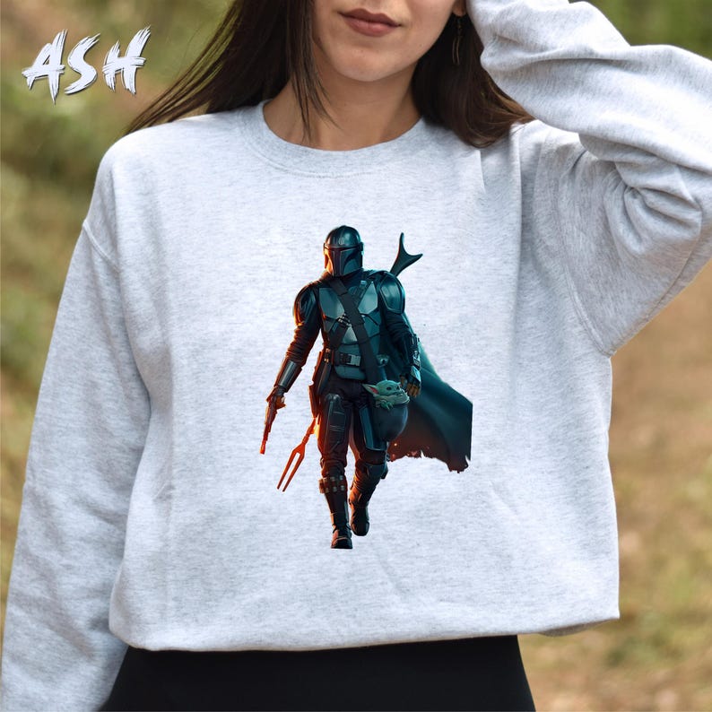May include: A grey sweatshirt featuring a printed image of a Mandalorian character from the Star Wars franchise. The character is wearing a helmet and armour and is holding a small, green creature.