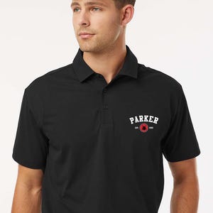 Puede incluir: Polo negro con cuello y mangas cortas. La camisa presenta el texto blanco "PARKER" sobre un emblema rojo. La palabra "black" est&aacute; en la esquina superior izquierda.
