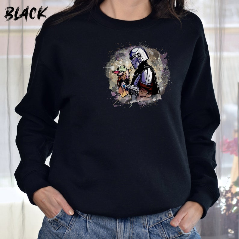 May include: A black sweatshirt with a watercolor-style image of a Mandalorian character holding a small, green creature. The image is centered on the front of the sweatshirt.