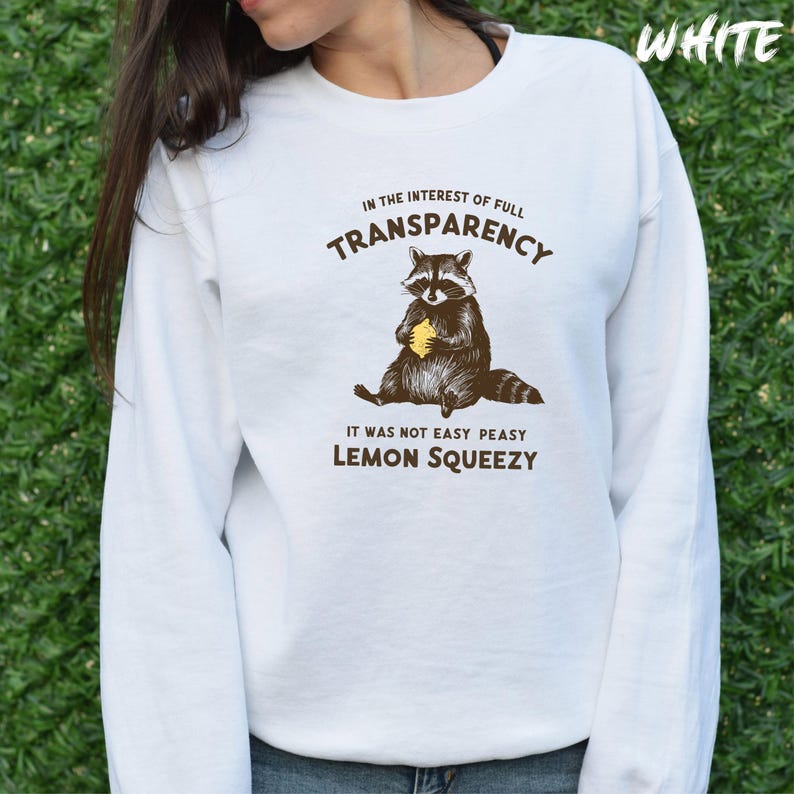 May include: White long-sleeved sweatshirt with a raccoon graphic holding a lemon. The text reads "In the interest of full transparency, it was not easy peasy lemon squeezy."