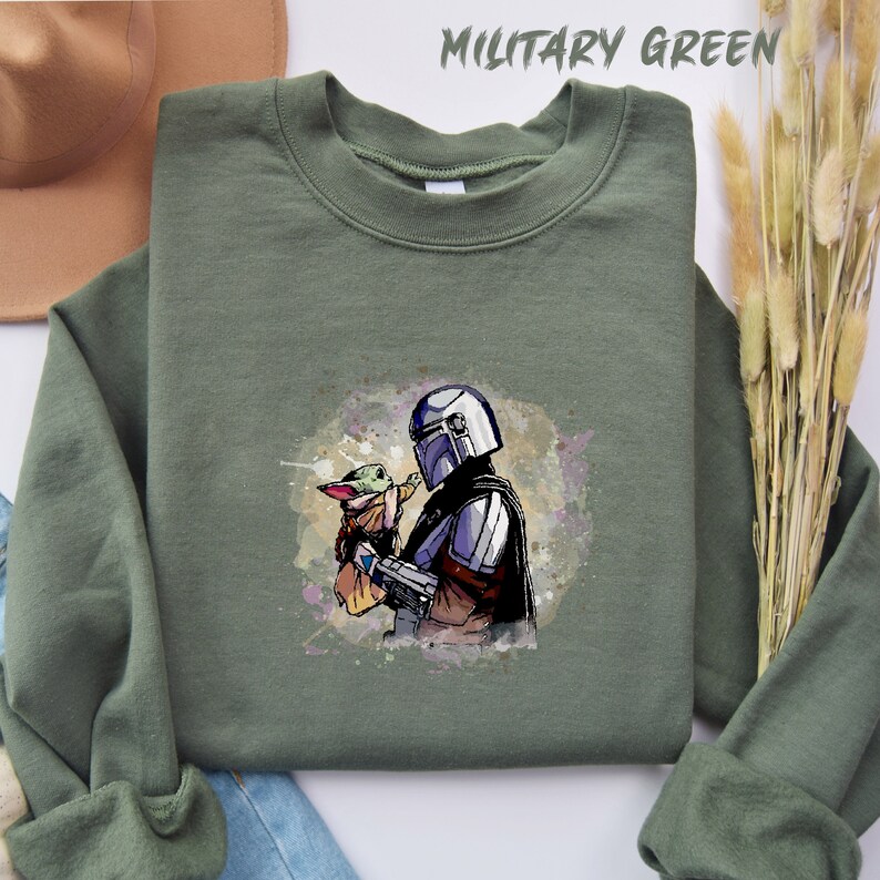 May include: A green sweatshirt with a watercolor-style image of a Mandalorian character holding a small, green alien creature. The image is centered on the sweatshirt.
