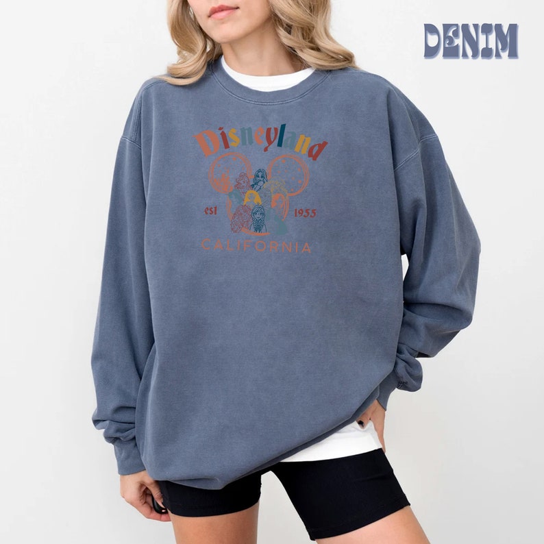 May include: A blue sweatshirt with a vintage-style Disneyland graphic. The graphic features the Disneyland logo, Mickey Mouse ears, and the text "est. 1955 California".