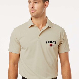 Puede incluir: Un polo color arena con mangas cortas y cuello. La camisa tiene la palabra "PARKER" en texto negro arqueado sobre un c&iacute;rculo rojo con un dise&ntilde;o. Se usa con pantalones grises oscuros y un cintur&oacute;n negro.