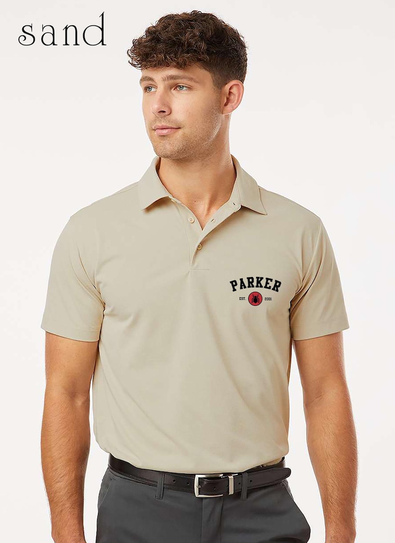 Puede incluir: Un polo color arena con mangas cortas y cuello. La camisa tiene la palabra "PARKER" en texto negro arqueado sobre un c&iacute;rculo rojo con un dise&ntilde;o. Se usa con pantalones grises oscuros y un cintur&oacute;n negro.