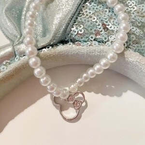 Könnte beinhalten: Ein weißes Perlenarmband mit einem silbernen Anhänger in Form eines Mäusekopfes mit Schleife. Der Anhänger hat kleine klare Strasssteine.