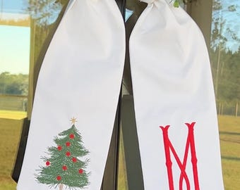 May include: White Christmas-themed decorative bow with a green embroidered Christmas tree on one side and a red embroidered letter "M" on the other. The bow is hanging in front of a window with greenery.