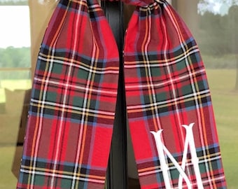 May include: A red, green, and blue plaid fabric ribbon hanging vertically with pointed ends. The ribbon features a white embroidered letter M monogram on the lower right side. The fabric has a woven texture with thin yellow and white accent lines.