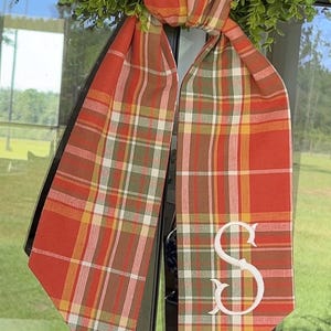 May include: A fabric ribbon featuring a plaid pattern in shades of orange, red, green, yellow, and white. The ribbon is tied in a knot at the top and hangs down in two long tails, with a large white embroidered letter S on the right tail.
