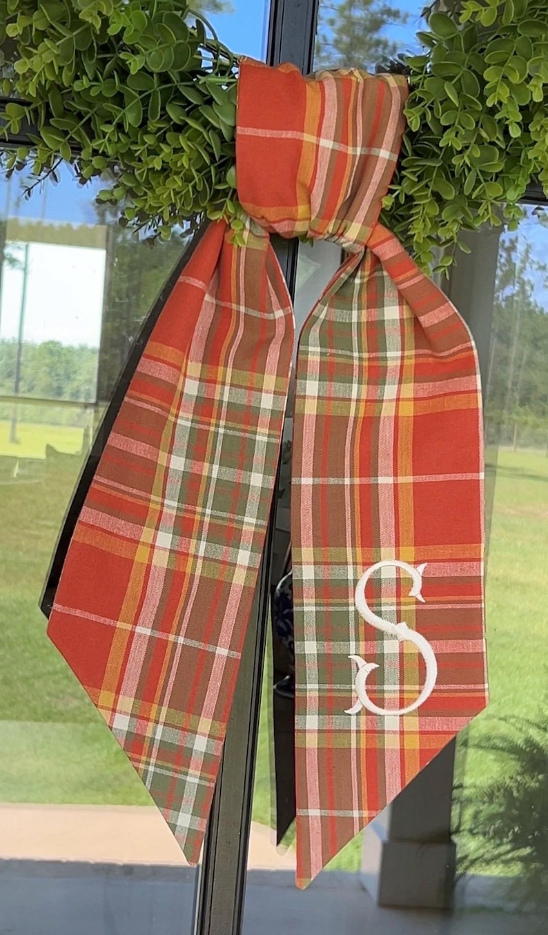 May include: A fabric ribbon featuring a plaid pattern in shades of orange, red, green, yellow, and white. The ribbon is tied in a knot at the top and hangs down in two long tails, with a large white embroidered letter S on the right tail.