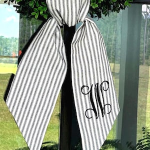 May include: A decorative bow with a black and white striped pattern, featuring a large, elegant monogram in black. The bow is tied around a dark structure, with a green wreath in the background. The design is suitable for home decor.