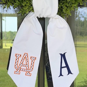 May include: A white fabric door hanger featuring two long, pointed tails. The left tail displays an orange and navy blue embroidered AU monogram with a cross-stitch pattern, while the right tail features a solid navy blue embroidered letter A.