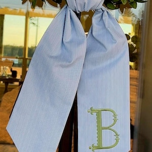 May include: Blue and white striped fabric bow with a green embroidered letter "B". The bow is tied around a green wreath. The fabric has a classic, preppy aesthetic.