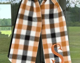 May include: A decorative scarf with a plaid pattern in white, black, and orange. The scarf is embroidered with the letter "S" in orange. The scarf is hanging in front of a window with a green wreath.