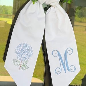 May include: White fabric bow with a blue hydrangea and the letter "M" embroidered on it.