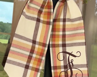 May include: A fabric ribbon featuring a plaid pattern in shades of cream, brown, orange, and yellow. The ribbon is tied in a knot and displays a large, dark brown embroidered monogram letter F in a cursive script on one of the hanging ends.